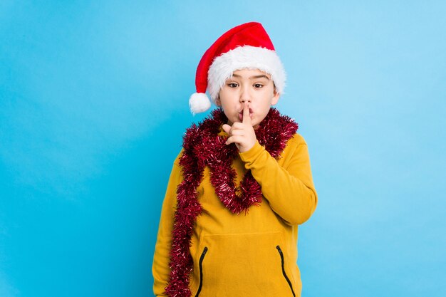 Niño celebrando el día de Navidad con un sombrero de santa aislado manteniendo un secreto o pidiendo silencio.