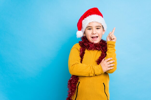 Niño celebrando el día de Navidad con un sombrero de santa aislado sonriendo alegremente señalando con el dedo lejos.