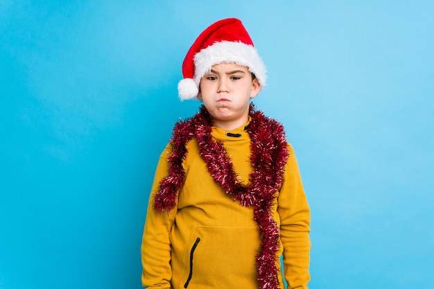 Niño celebrando el día de Navidad con un sombrero de santa aislado sopla mejillas, tiene expresión cansada. Concepto de expresión facial