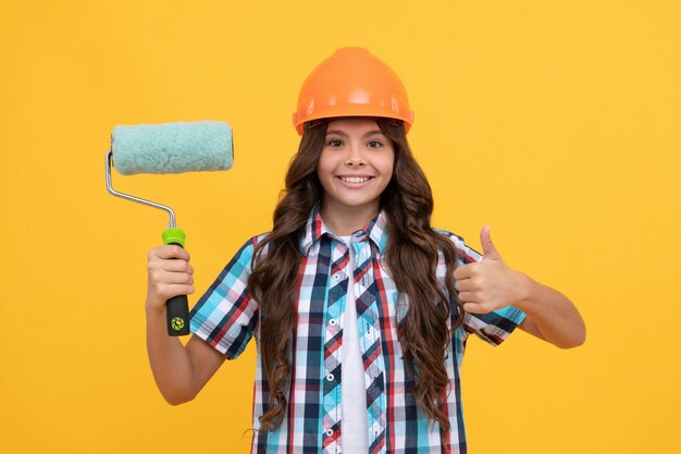 Niño en cepillo de rodillo de casco para decoración trabajo de renovación de educación infantil mejorar su infancia futuro ingeniero mejora pulgar arriba haciendo reparación de superficie niña adolescente feliz sostener rodillo de pintura