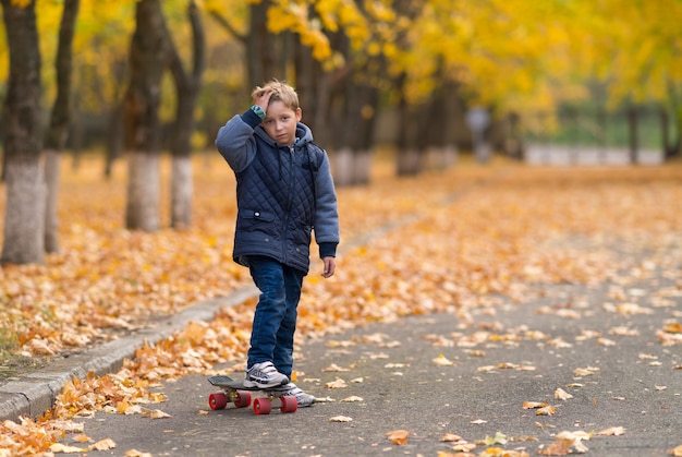 Niño en chaqueta abrigada con patineta sosteniendo su mano en la frente, como si estuviera herido, de pie solo en el parque. Árboles y hojas amarillas