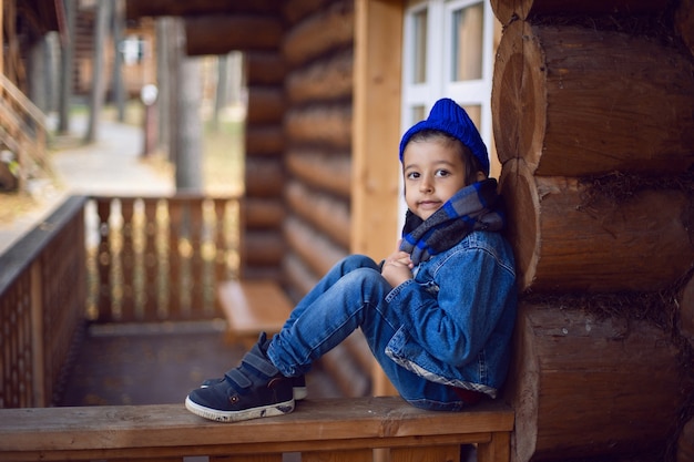 Niño en una chaqueta de mezclilla azul y un sombrero está sentado en una casa de madera en otoño