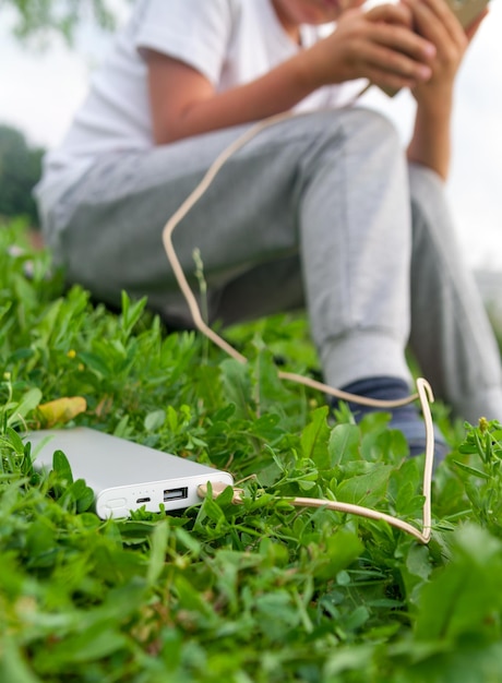 Niño chateando en un teléfono inteligente en un césped mientras se carga desde el banco de energía