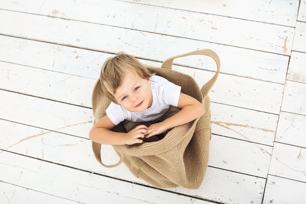 Niño chico hermoso lindo, alegre, divertido y feliz en una bolsa de mimbre sobre un piso blanco de madera