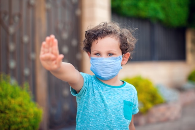 Niño chico con mascarilla médica mostrando gesto de parada al aire libre