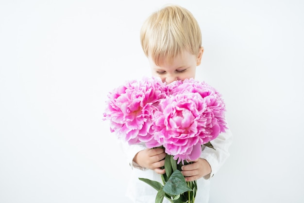 Niño chico rubio con gran ramo de peonías rosas sobre fondo blanco amor y concepto romántico