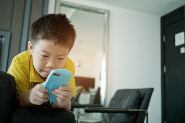 Niño chino asiático jugando teléfono inteligente en la cama, el niño usa el teléfono y juega, adictos y dibujos animados,