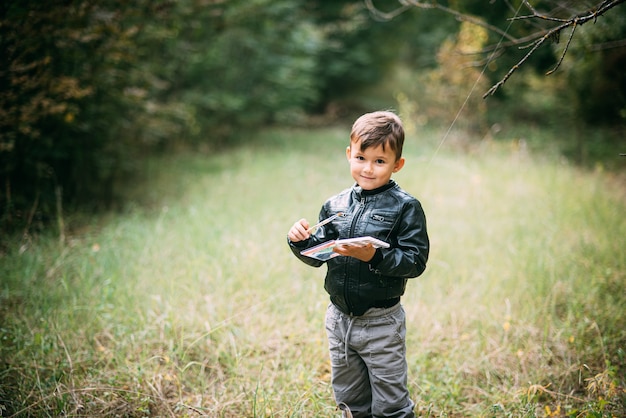 Un niño de cinco años está parado en el bosque con pinturas en sus manos, tratando de pintar las hojas de los árboles con pinturas.