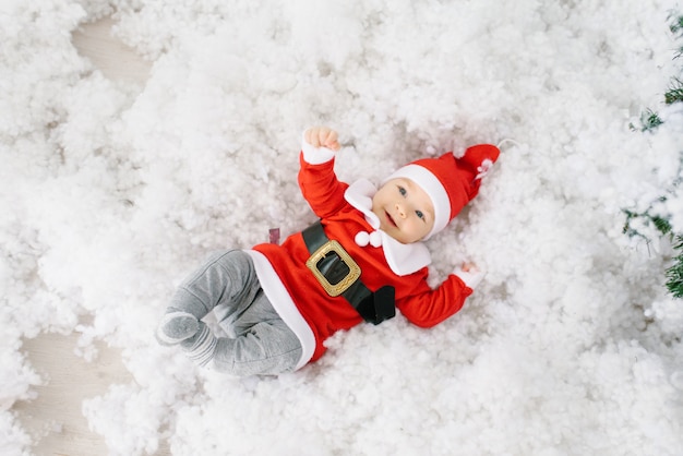 Un niño de cinco meses con un traje de Papá Noel está acostado en la nieve artificial sobre su espalda.