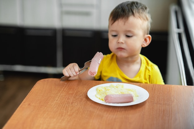 Foto niño en la cocina comiendo salchichas y puré de papas