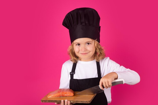 Niño cocinero sostenga la tabla de cortar con bistec de salmón de pescado y cuchillo Kid boy en sombrero de chef y delantal sobre fondo rosa estudio aislado