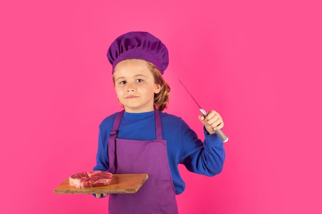 Foto niño cocinero sostiene la tabla de cortar con carne de res y cuchillo niño con uniforme de cocinero y sombrero de chef preparando comida en el retrato del estudio de cocina concepto culinario y de comida para niños
