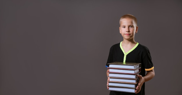 Niño colegial en camiseta negra sosteniendo una pila de libros sobre una pared gris