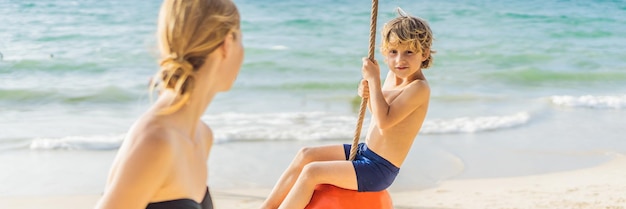 Un niño en un columpio en la playa, mamá e hijo pasan tiempo en la pancarta de playa de formato largo