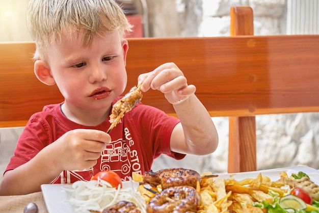 El niño come carne con patatas fritas y verduras en la terraza del restaurante al aire libre