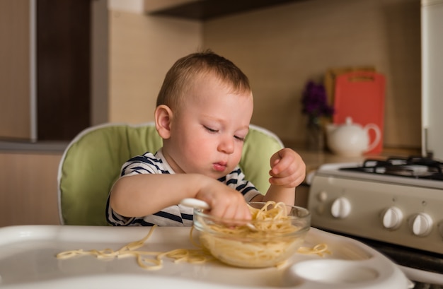 Un niño come espagueti en una mesa alta de la cocina. Aprendiendo a comer de forma independiente.