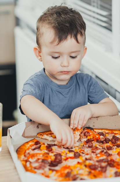 El niño se come una pizza enorme y dañina él mismo en la cocina y bebe jugo, muy graso y dañino.