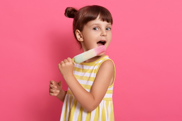 Niño comiendo helado mientras posa aislado en rosa, vestido de verano con rayas blancas y amarillas