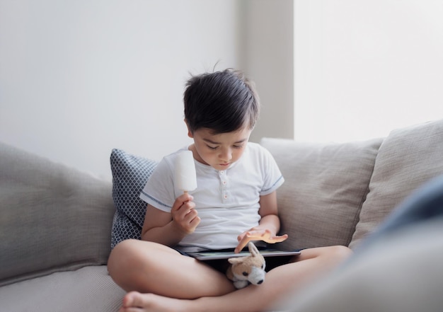 Niño comiendo una piruleta de hielo de coco mientras juega un juego en una tabletaRetrato de tecla altaNiño sentado en el sofá haciendo la tarea en línea en casaUn niño jugando juegos en Internet con la luz que brilla por la ventana