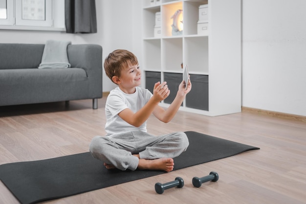 El niño se comunica en un teléfono inteligente mientras hace fitness en casa en la sala de estar.