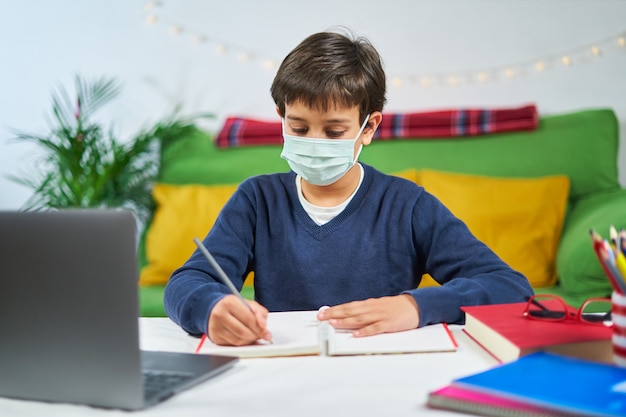 Niño concentrado con mascarilla escribiendo en la computadora portátil, interior de la casa, espacio libre