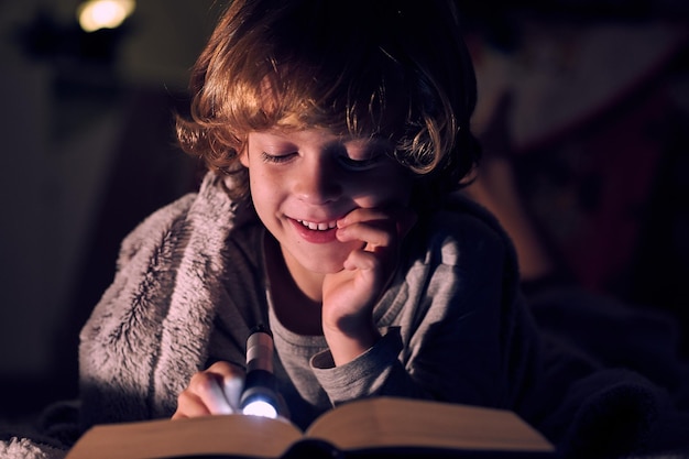 Un niño contento leyendo un libro interesante con una linterna brillante mientras descansa en la cama bajo una manta en una habitación oscura en casa