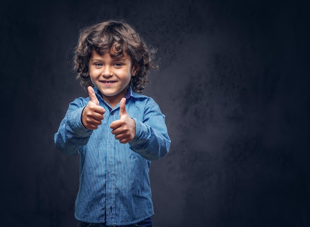 Niño contento lindo colegial con cabello castaño rizado vestido con una camisa azul, mostrando los pulgares hacia arriba en un estudio.