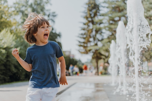 Un niño corre por la calle y juega con los chorros de agua de una fuente que brota del suelo. niño con camiseta azul y pantalones cortos blancos está jugando al aire libre. estilo de vida. espacio para texto