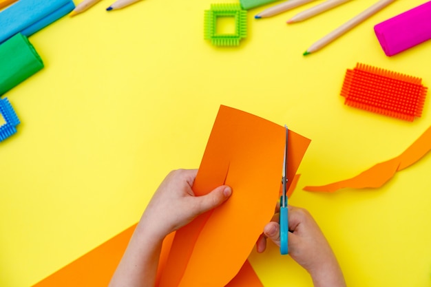 Niño cortando papel naranja de color con unas tijeras en una mesa para hacer algunos trabajos manuales