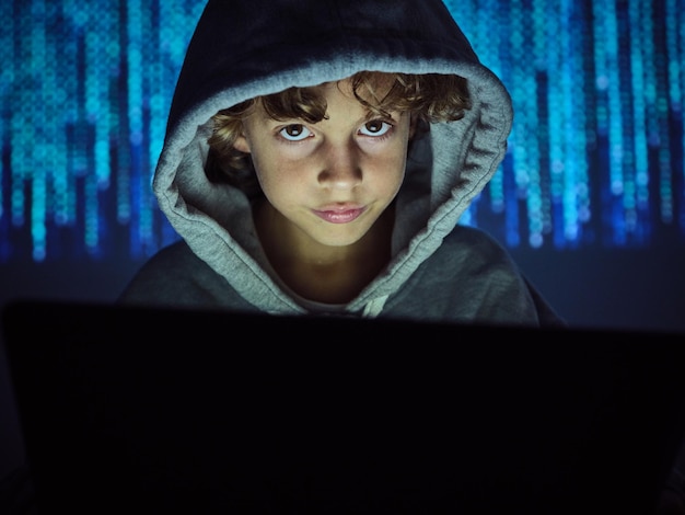 Foto niño de cosecha sentado en una computadora portátil y pirateando mientras mira la cámara en un fondo de código binario borroso en luz oscura