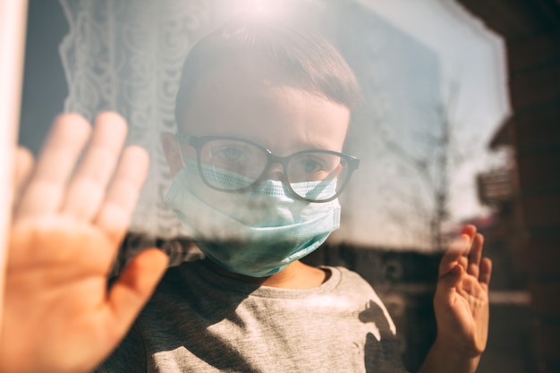 Un niño en cuarentena debido a un virus se sienta en su casa con una máscara y mira por la ventana en un día soleado. Concepto de coronavirus y contaminación del aire pm2.5. COVID-19