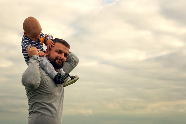 Un niño en el cuello de su padre. Camina cerca del agua. Bebé y papá contra el cielo.