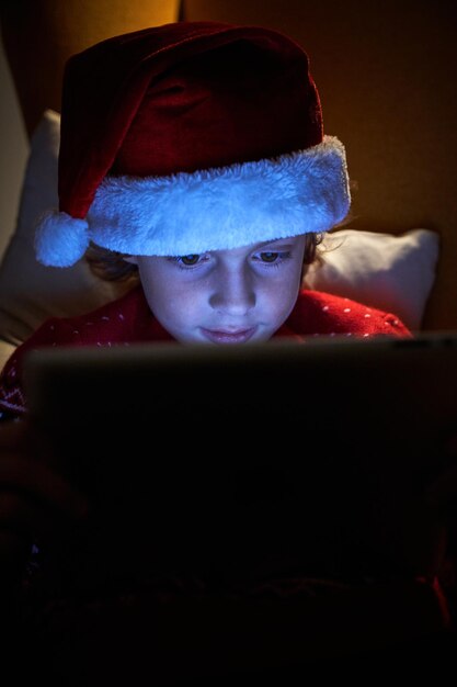 Un niño curioso con gorro de Papá Noel usando una tableta moderna mientras se sienta en un cómodo sillón en casa por la noche