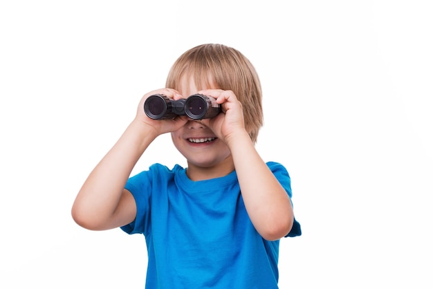 Niño curioso. Niño alegre mirando a través de binoculares y sonriendo mientras está de pie aislado en blanco