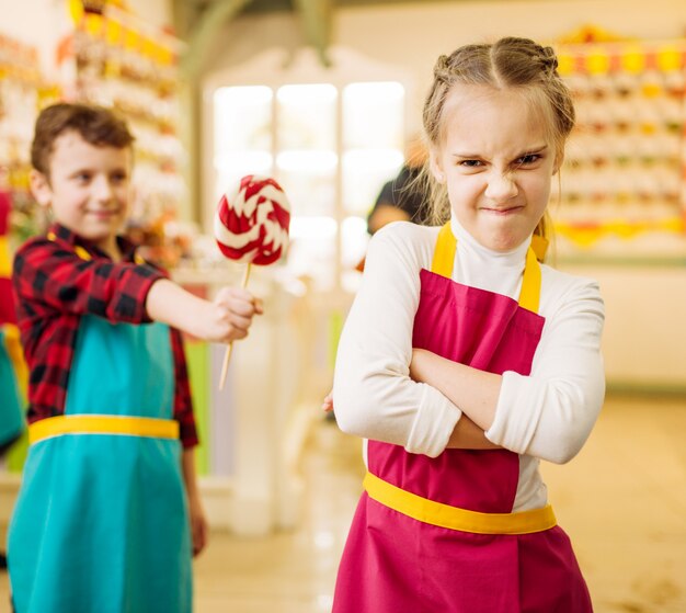 Niño da piruleta hecha a mano a niña obstinada. Niños en taller en pastelería. Diversión navideña en la tienda de dulces. Caramelo de azúcar cocido fresco