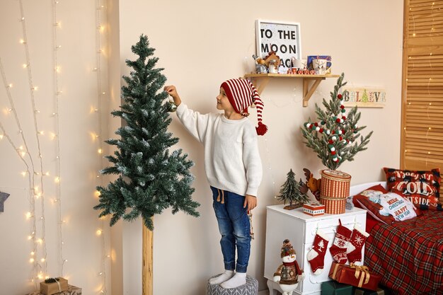 Niño decorando el árbol de Navidad en casa. Niño con sombrero rojo con adorno de Navidad. Los niños celebran las vacaciones de invierno. Los niños decoran la sala de estar para Navidad.