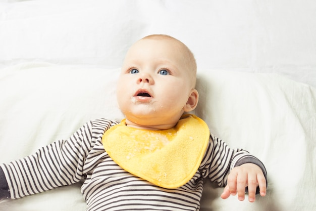 Niño con delantal amarillo para niños con residuos de comida se sienta en una cama blanca después de comer.