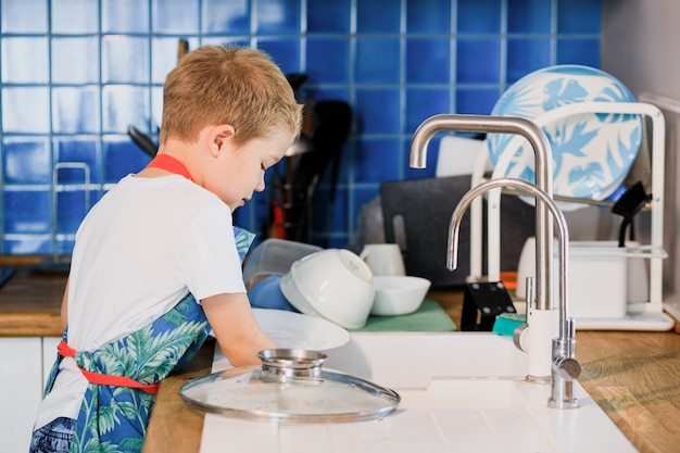 Un niño con un delantal lava platos en la cocina de casa