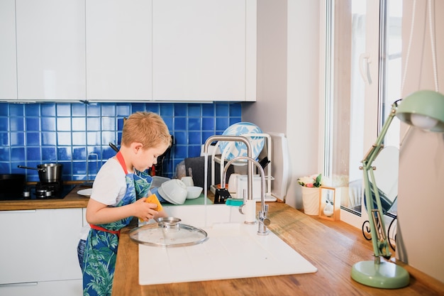 Un niño con un delantal lava platos en la cocina de casa