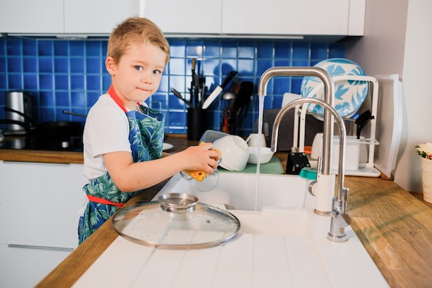 Un niño con un delantal lava platos en la cocina de casa