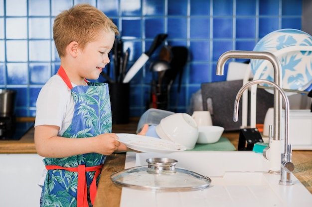 Un niño con delantal lava platos en la cocina de su casa.