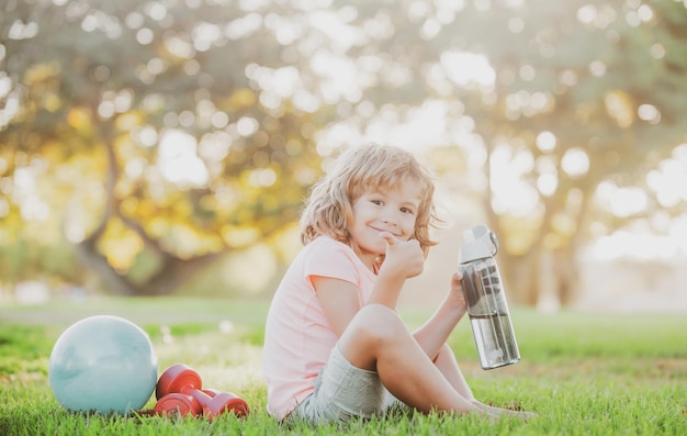 Niño deportivo bebiendo agua en el parque Concepto deportivo para niños Ejercicios de salud y energía para niños con pesas Estilo de vida saludable para niños