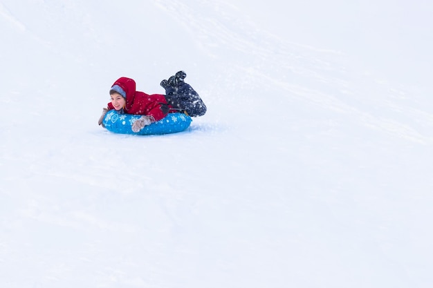 Niño deslizándose sobre un tubo azul con ropa de invierno entre la nieve blanca. Paseos en trineos hinchables.