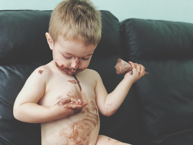 Foto niño desordenado sin camisa comiendo helado mientras está sentado en el sofá en casa