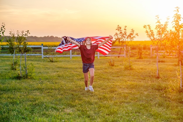 Niño despreocupado corriendo en el campo con la bandera americana soplando en las manos al atardecer