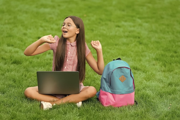 Un niño despreocupado se relaja en el parque sobre hierba verde trabajando en una computadora para la educación en línea con la educación de la mochila escolar