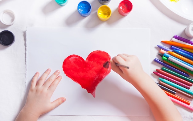 Un niño dibuja un corazón rojo con pinturas de colores en el papel.