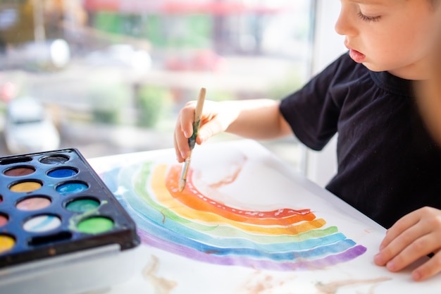 El niño dibuja con pinturas multicolores sobre papel un arco iris sentado en una mesa en casa una vista de cerca ...