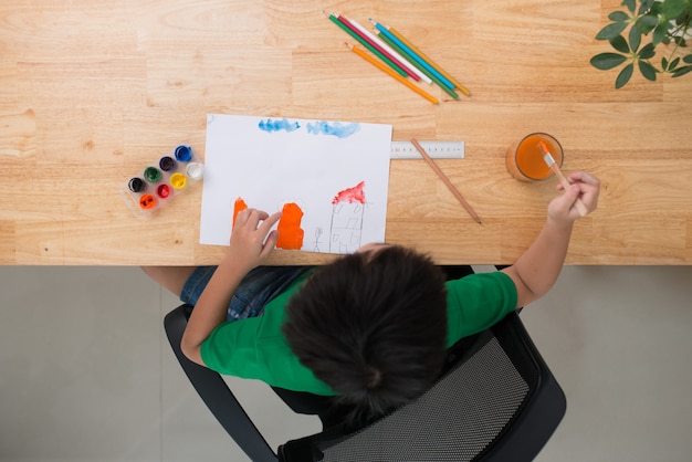 Niño dibujando en el papel. Niño sentado en la silla a la mesa con pinturas