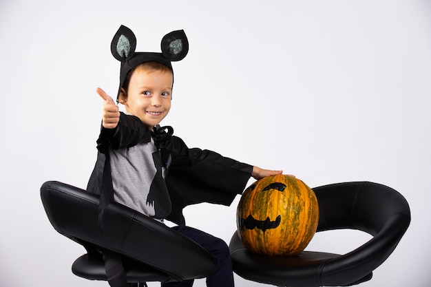 Un niño disfrazado de murciélago se sienta en sillas negras con una calabaza. Truco o trato, composición de halloween.